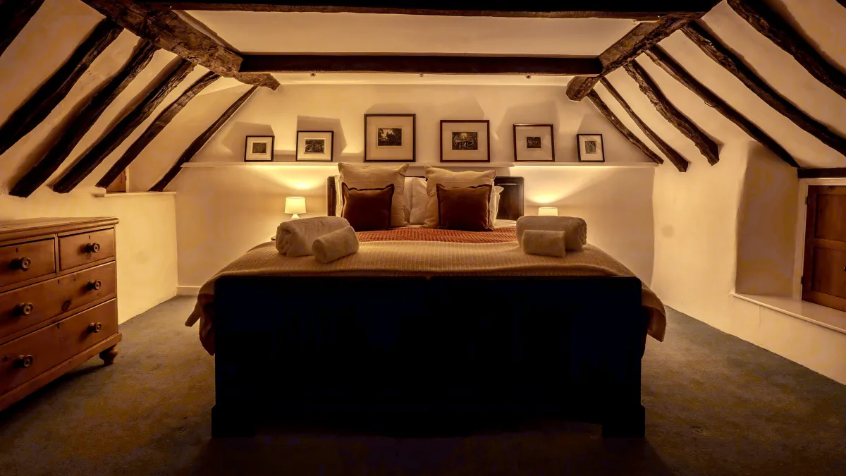 A low, lamplit view of the super king bedroom showing the full sweep of oak beams and the row of framed prints lit above the headboard