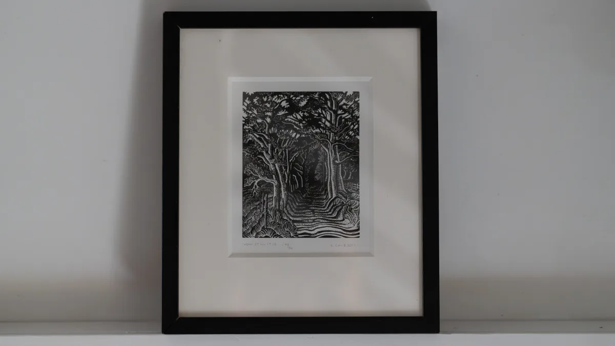 A black-framed wood engraving of trees arching over a country lane, propped on the white plaster ledge