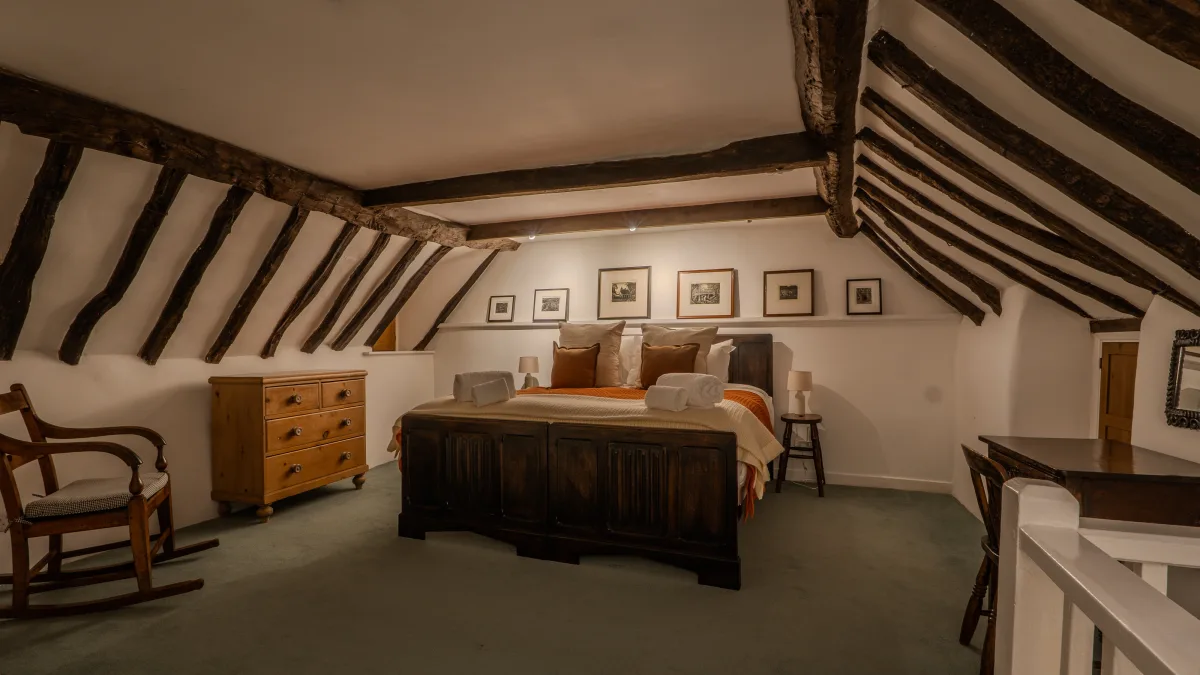 The full sweep of the room with the wooden rocking chair in the near corner, the pine dresser and bed set beneath the fan of oak beams